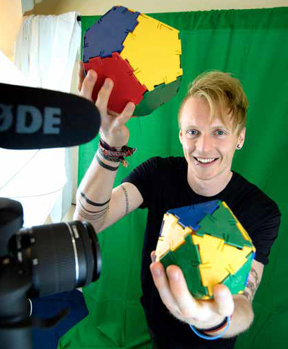 Tm Crawford holding multi-coloured three dimensional shapes in front of a camera, with a green-screen behind him