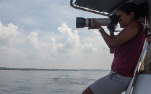 Asha de Vos stood on a boat, using a camera 