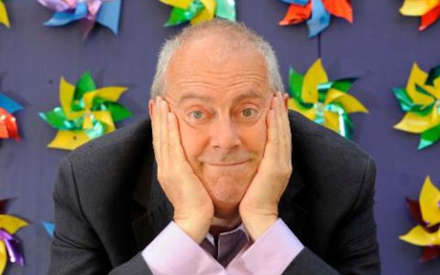 Gyles Brandreth, with his chin on his hands, in front of a wall of colourful windmills