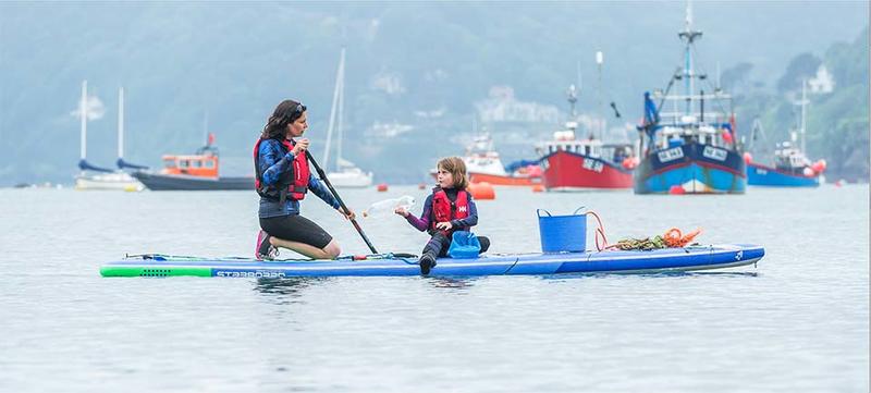 A mother and daughter sat on a paddleboard at sea, with buckets and litter on the paddleboard, and boats in the background