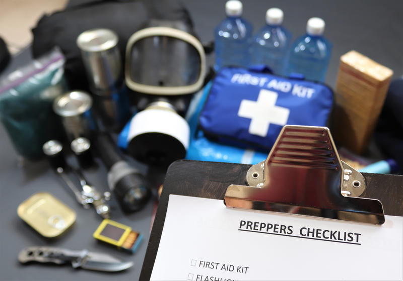 In the foreground is a clipboard with a title 'Preppers checklist', and in the background are various items including medical supplies, water, torch and knife